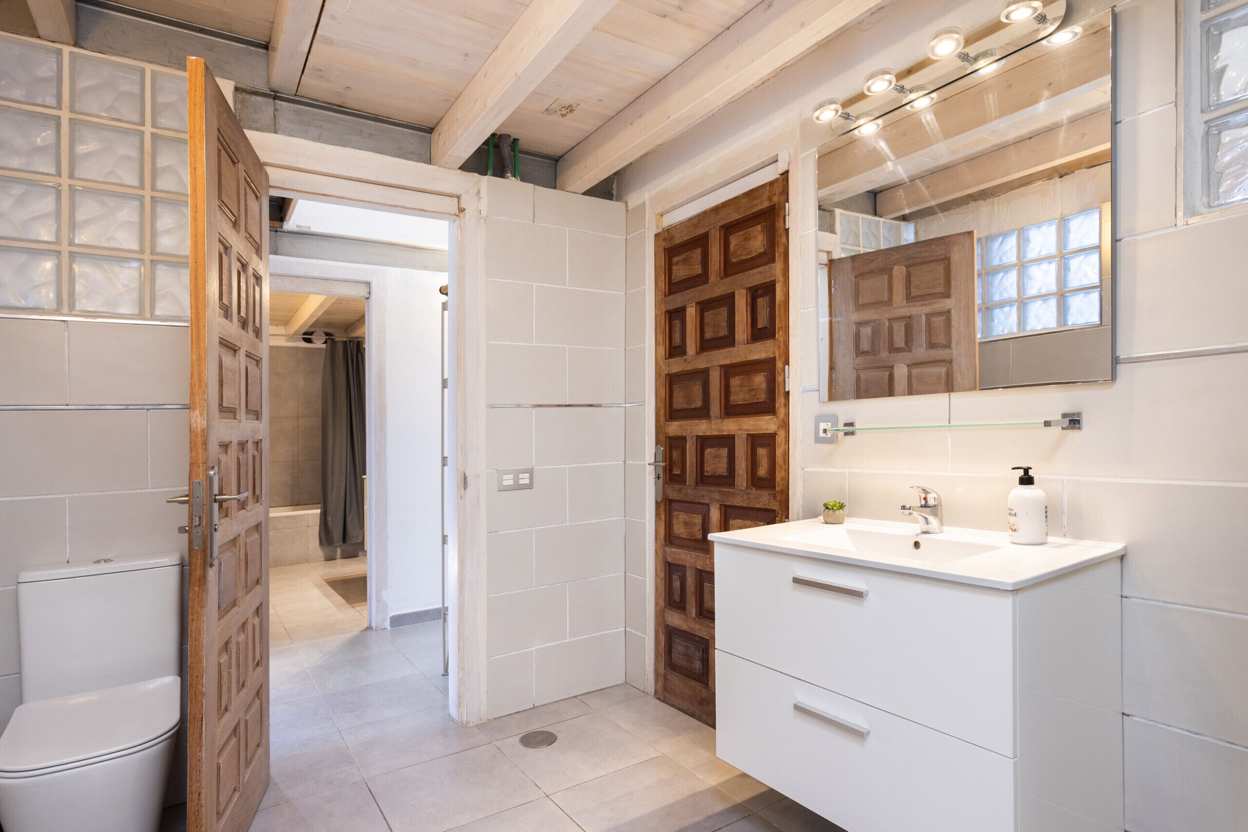 A modern bathroom with a wooden door, white tiles, a white sink, and a mirror with lights.