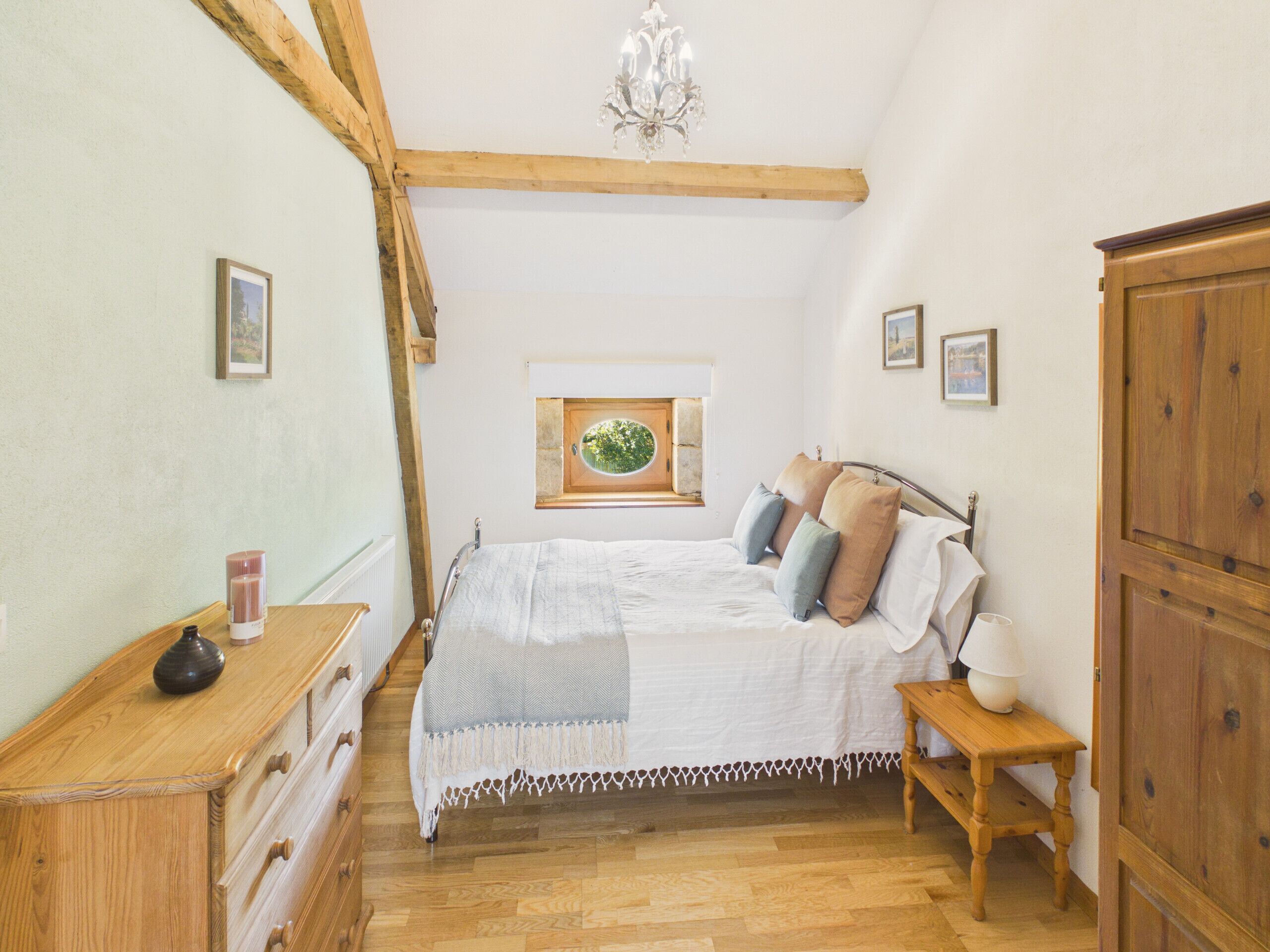 A bedroom with a wooden bed, a nightstand with a lamp, a dresser, a window with a view, and a chandelier.