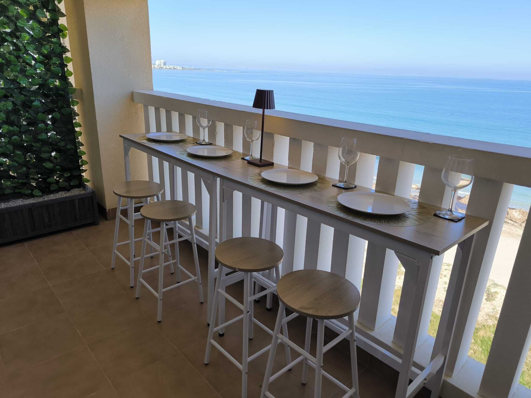 Un balcon avec vue sur l’océan, une table dressée avec des assiettes et des verres, et des tabourets de bar blancs.