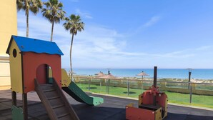 Un parque infantil con tobogán y estructura para escalar, con vistas a una playa con sombrillas y palmeras.