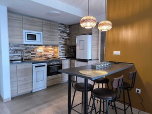 Eine moderne Küche mit Mikrowelle, Herd und Kühlschrank. Zwei Pendelleuchten, ein Esstisch mit Hocker und eine strukturierte Spritzschutzwand.