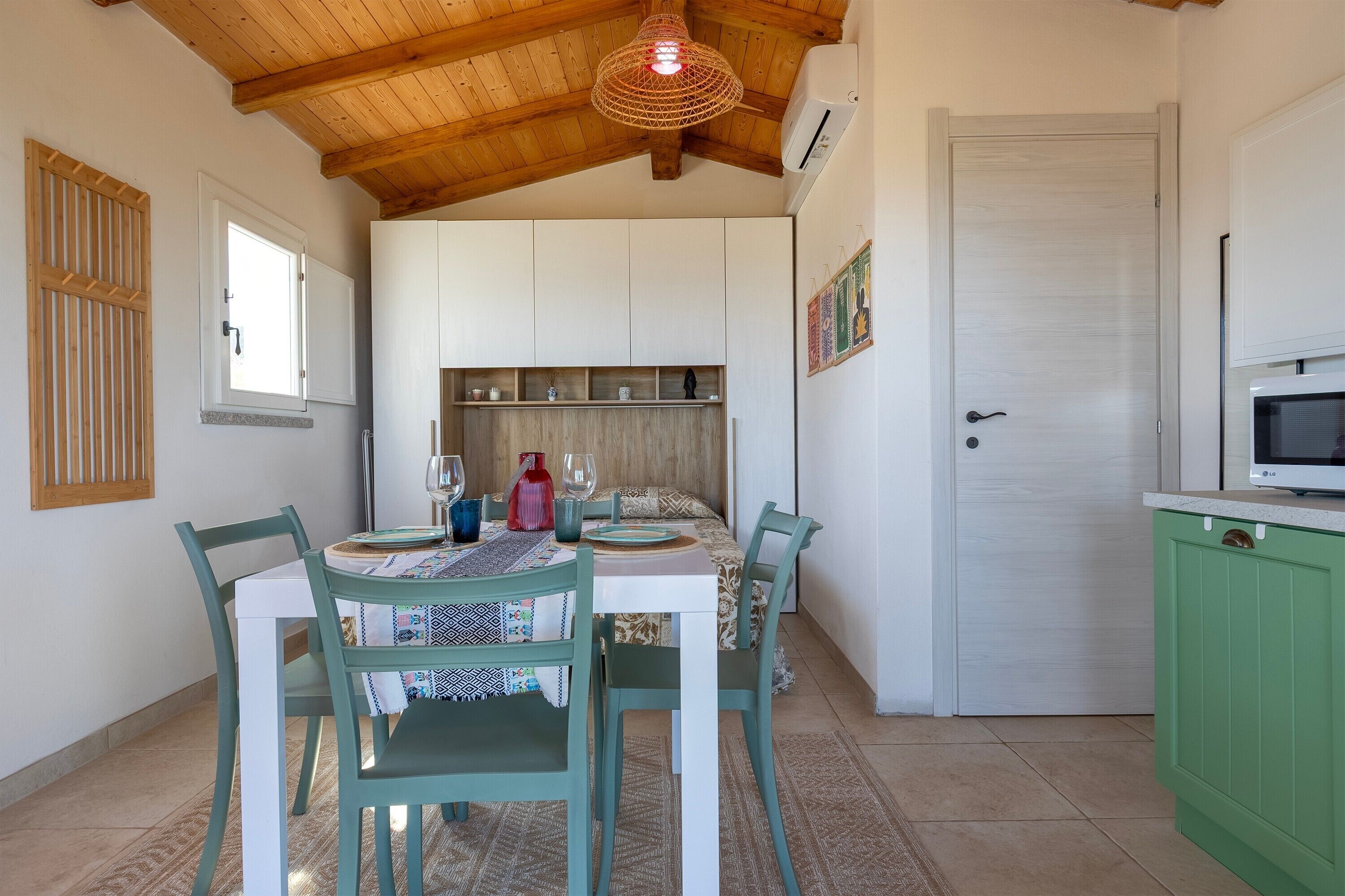 A dining area with a table set for four, chairs, a hanging light fixture, and a kitchen area with cabinets and a microwave.