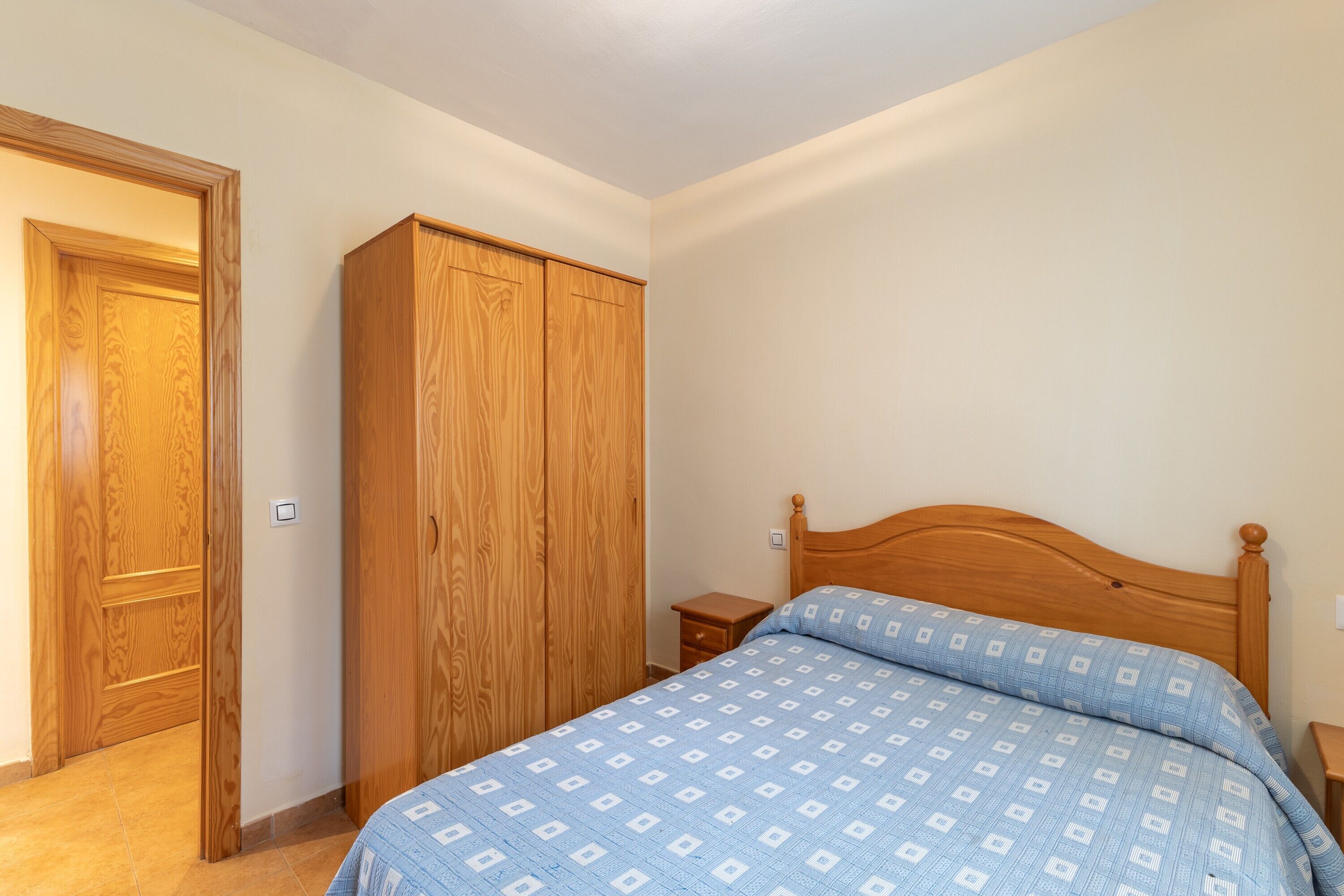 A bedroom with a wooden wardrobe, a single bed with a patterned bedspread, and a nightstand.