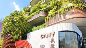 En moderne bygning med en buet facade, vertikale trælameller og grønne planter på altanerne. Navnet "CANYAS" er tydeligt vist på bygningen.