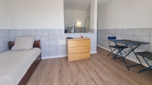 Una habitación pequeña con una cama, un pequeño aparador de madera, una mesa plegable y una kitchenette al fondo.
