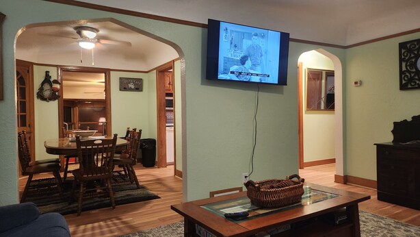 A living room with a television mounted on the wall, a dining area with wooden furniture, and a kitchen visible through an open doorway. - Charming 2-bedroom apartment with AC in vibrant Austin,Mn (Austin)