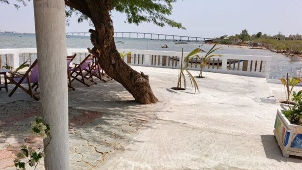 A paved outdoor area with a tree, chairs, and a railing overlooking a body of water with a bridge in the distance. - A La Teranga (Foundiougne)