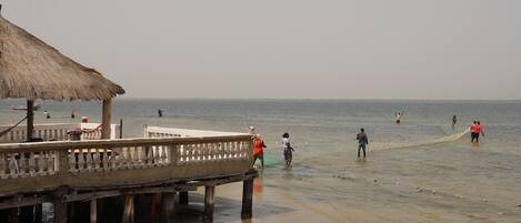 A beach with a thatched-roof structure, a pier, and people fishing in the water.