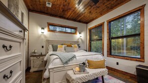 A bedroom with a wooden ceiling, a bed with multiple pillows, a bedside table with a bottle and glasses, and two windows with blinds. - 2BR Family Retreat with Bunk Room and Hot Tub (Broken Bow)