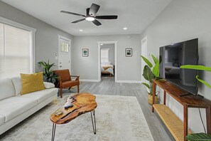 Ein Wohnzimmer mit einer Couch, Couchtisch, Fernseher, Deckenventilator und einem Flur, der zum Schlafzimmer führt.