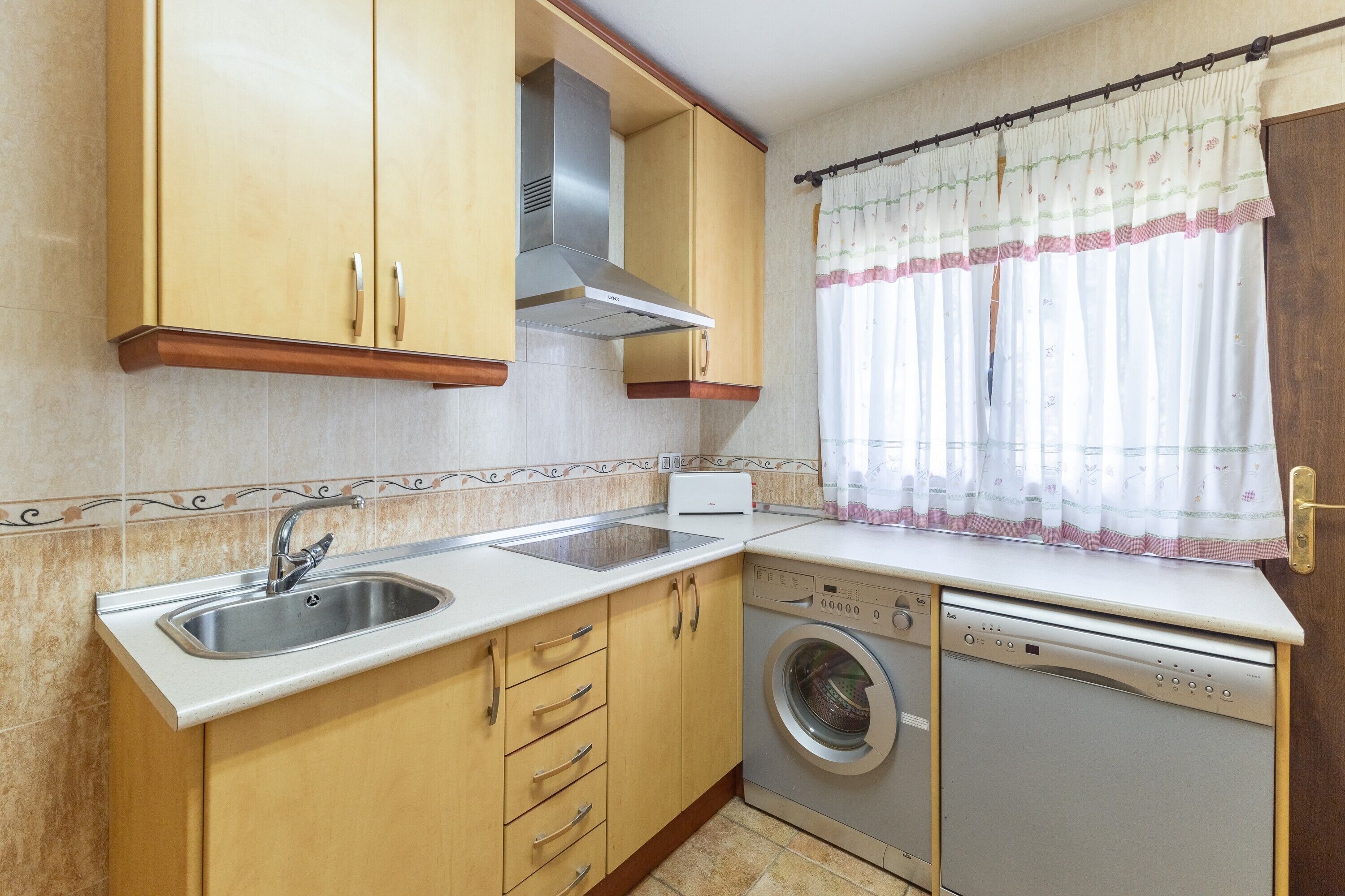 A compact kitchen with a sink, a built-in washing machine, wooden cabinets, and a window with a curtain.
