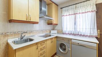 A compact kitchen with a sink, a built-in washing machine, wooden cabinets, and a window with a curtain.