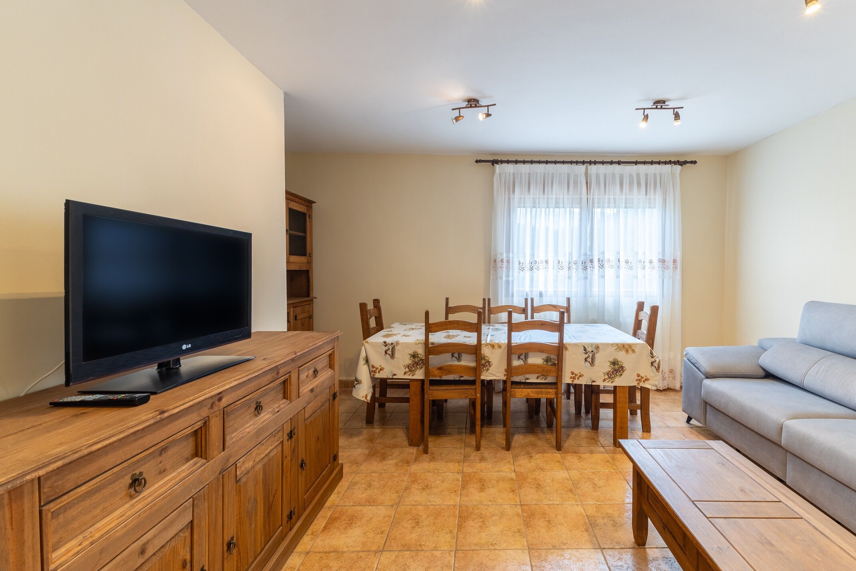 A living room with a TV on a wooden cabinet, a dining table with chairs, a sofa, and a window with curtains.