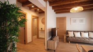 Ein modernes Wohnzimmer mit Holzboden, einer Couch mit Kissen, einem Fernseher auf einem Holzschrank und einer Pflanze in der Ecke.