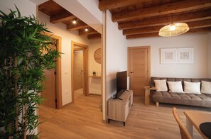 Ein modernes Wohnzimmer mit Holzboden, einer Couch mit Kissen, einem Fernseher auf einem Holzschrank und einer Pflanze in der Ecke.