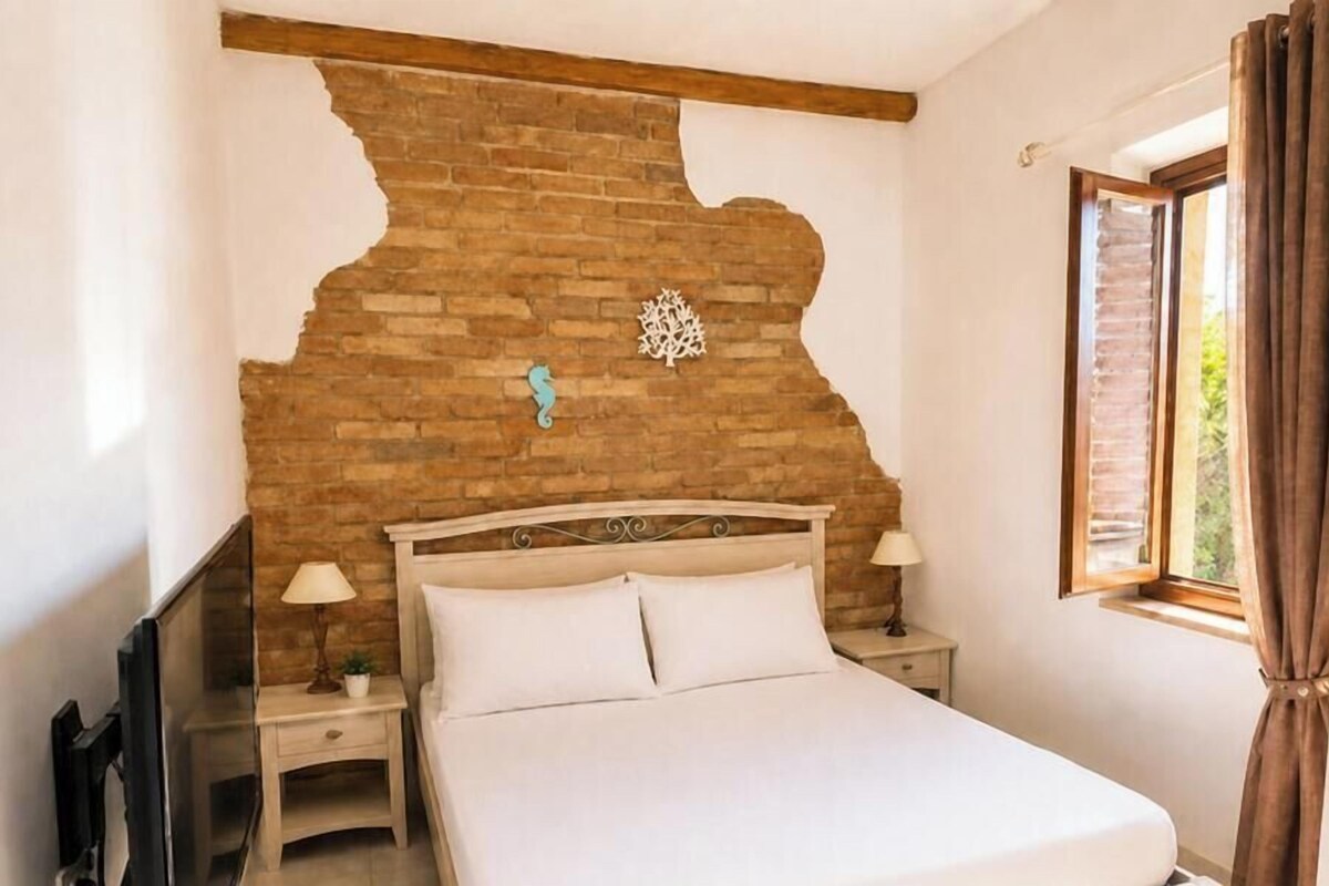Un dormitorio con pared de ladrillo, una cama, dos mesitas de noche con lámparas y una ventana con cortinas.