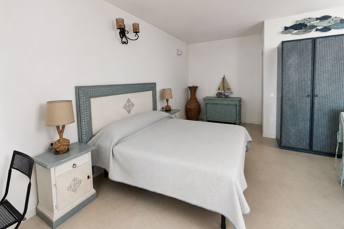 Un dormitorio con cama, mesitas de noche, una silla, cómoda, armario y lámparas de pared.
