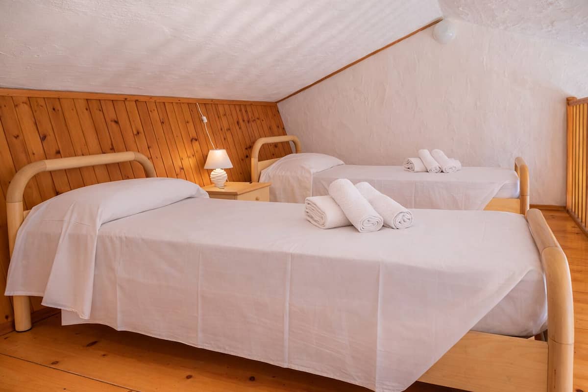 Una habitación con dos camas de madera, cada una con una colcha blanca y toallas enrolladas. Hay una mesita de noche con una lámpara entre las camas.