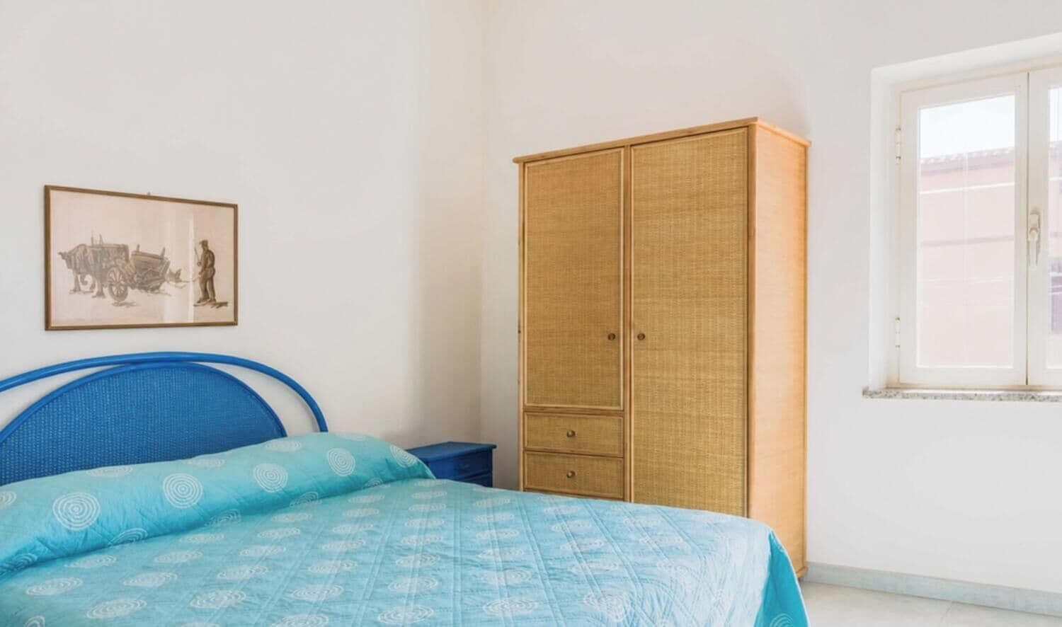 Een slaapkamer met een bed, een kledingkast, een nachtkastje, een ingelijste foto aan de muur en een raam.