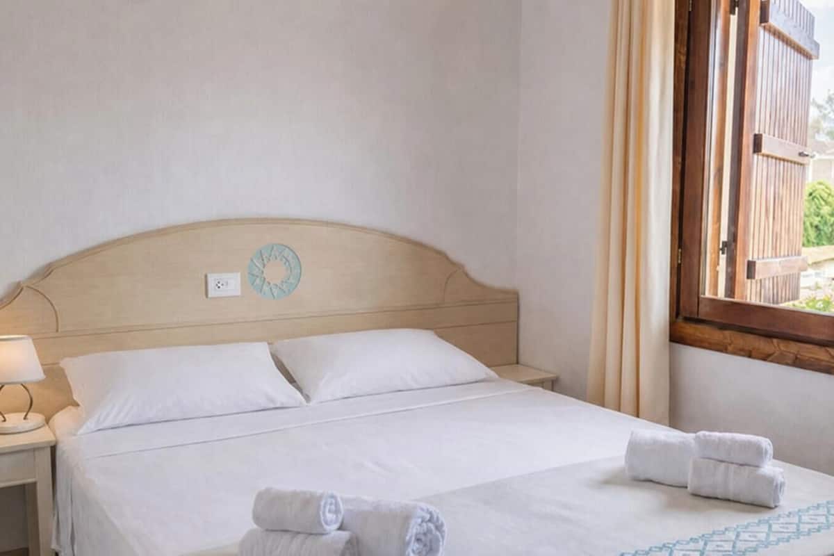 Una habitación de hotel con cama, una mesita de noche con lámpara, una ventana con cortinas y vistas al exterior.