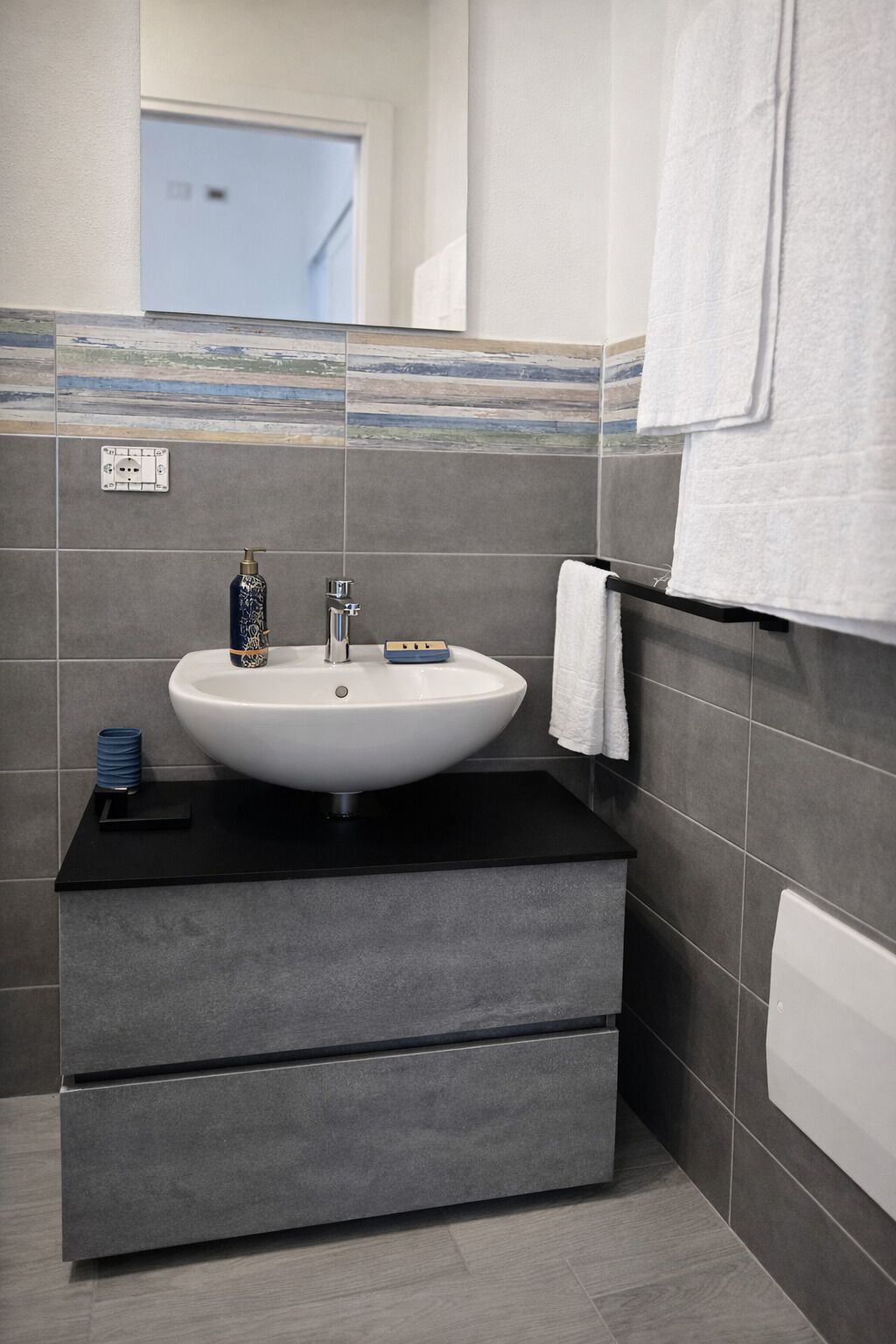 A bathroom with a white sink, dark tiles, a towel rack, and a door in the background.
