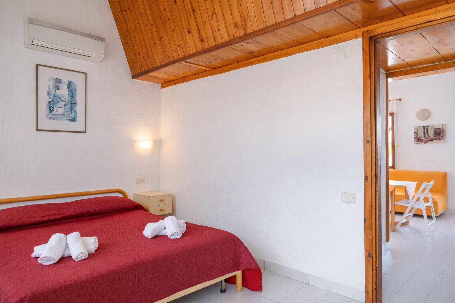 Ein Schlafzimmer mit Holzbalkendecke, einem Bett mit roter Bettwäsche, einem Nachttisch, einer Wandleuchte und einem gerahmten Bild.