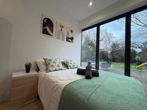 A bedroom with a bed, pillows, a nightstand, a small plant, and a large window with a view of the outdoors.