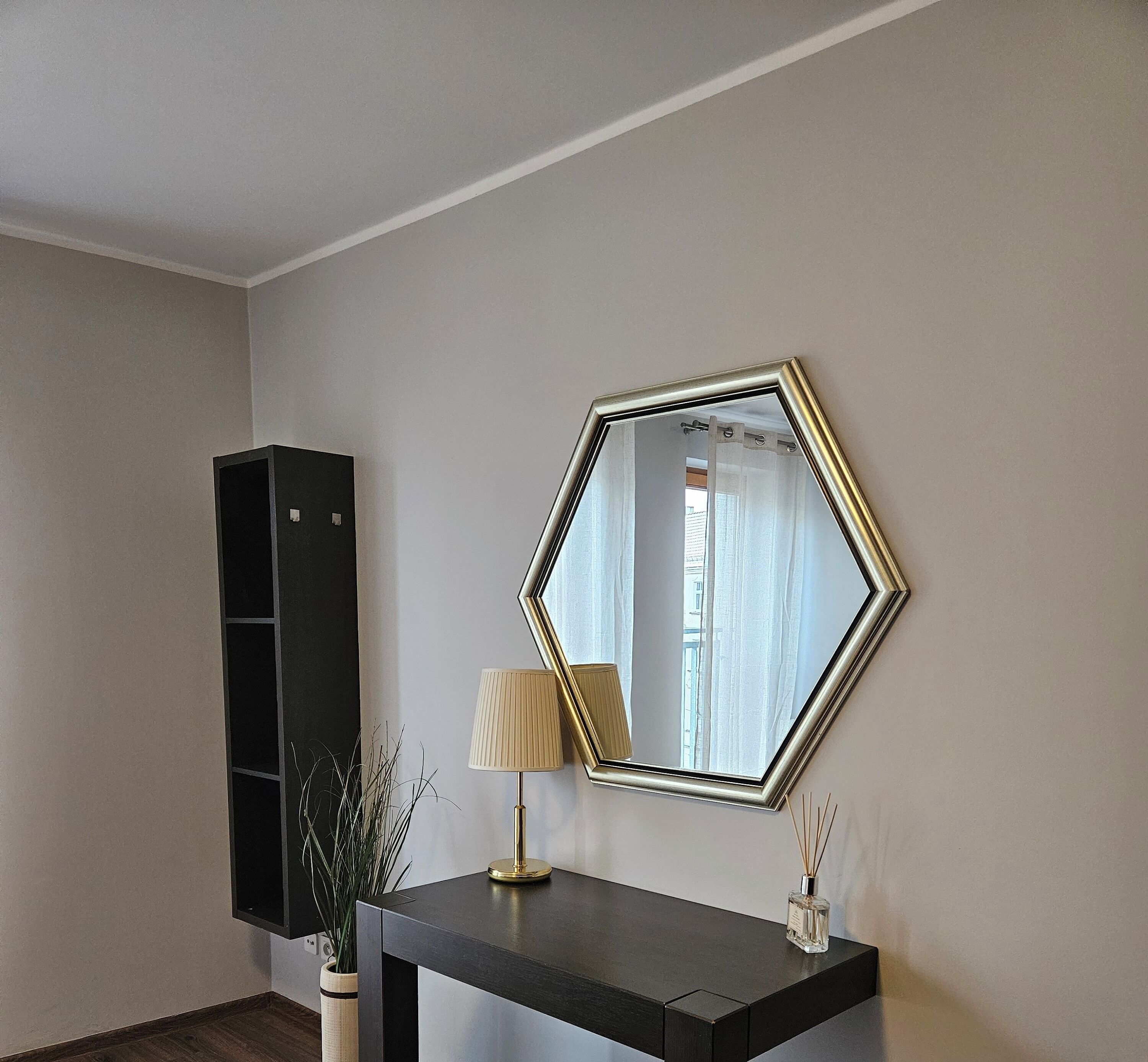 Una habitación con un espejo adosado a la pared, una consola oscura, una lámpara, una planta en maceta y un estante negro alto.