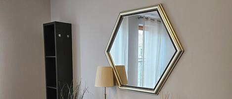 Una habitación con un espejo adosado a la pared, una consola oscura, una lámpara, una planta en maceta y un estante negro alto.