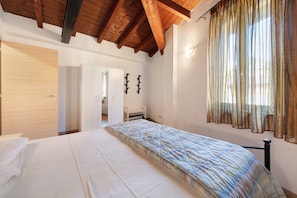 Ein Schlafzimmer mit Bett, Holzbalkendecke, Kleiderschrank, Schreibtisch und einem Fenster mit Vorhängen.