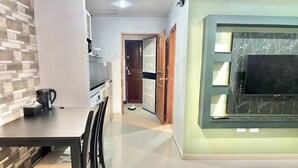A modern kitchen with a coffee maker, a dining area with chairs, a TV mounted on a wall, and a door leading to another room. - Large Condo Middle of Jomtien, Near the Beach 42m2 (Pattaya)