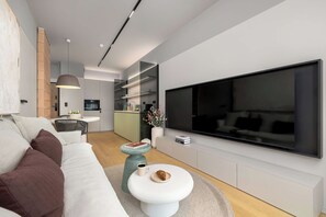 Een moderne woonkamer met een grote flatscreen televisie, een witte bank met bruine kussens, een eethoek met een tafel en stoelen, en een keuken op de achtergrond.