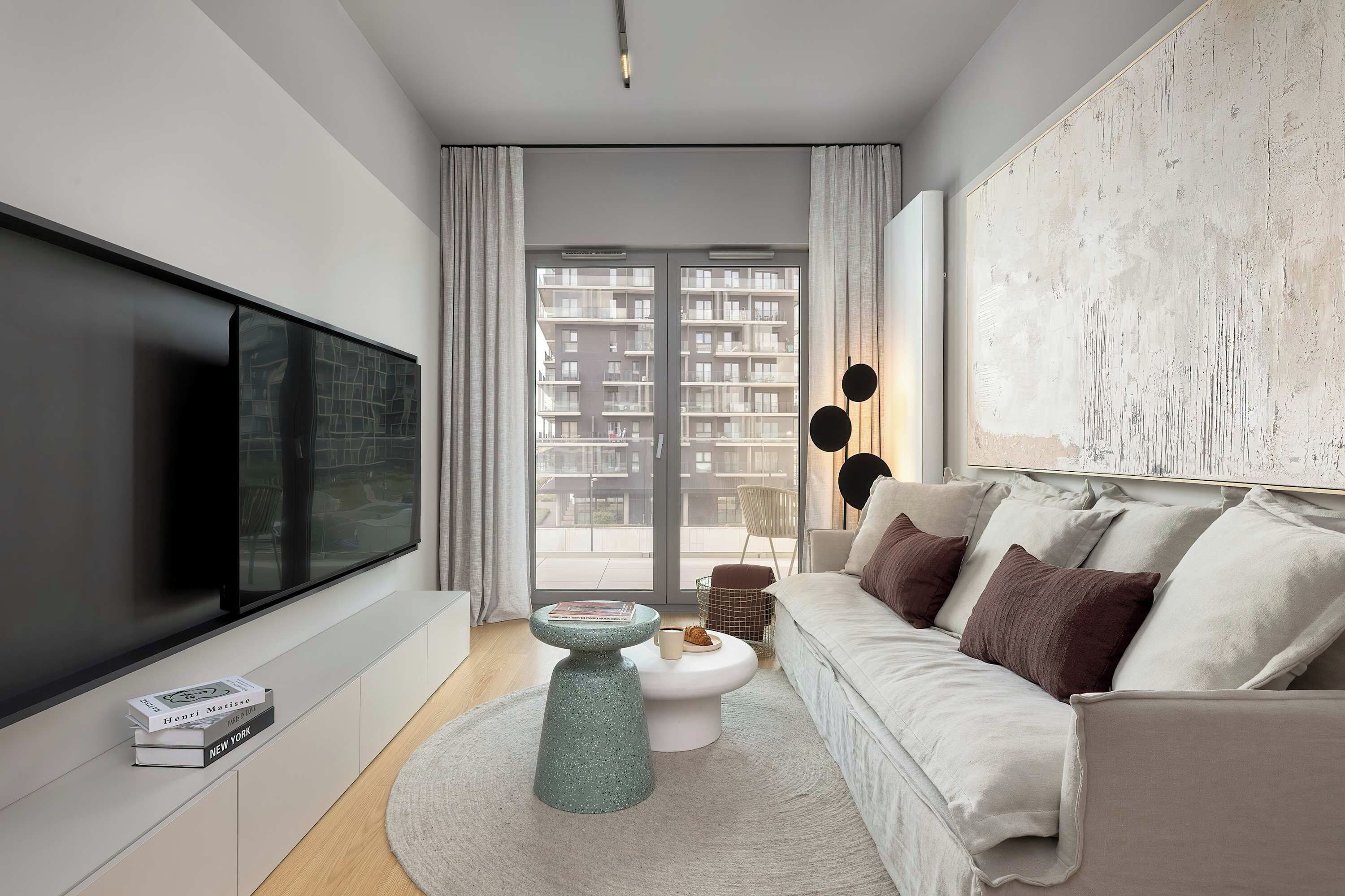 A modern living room with a large flat-screen TV, a round coffee table, a sofa with cushions, and a view of a residential building through the window.
