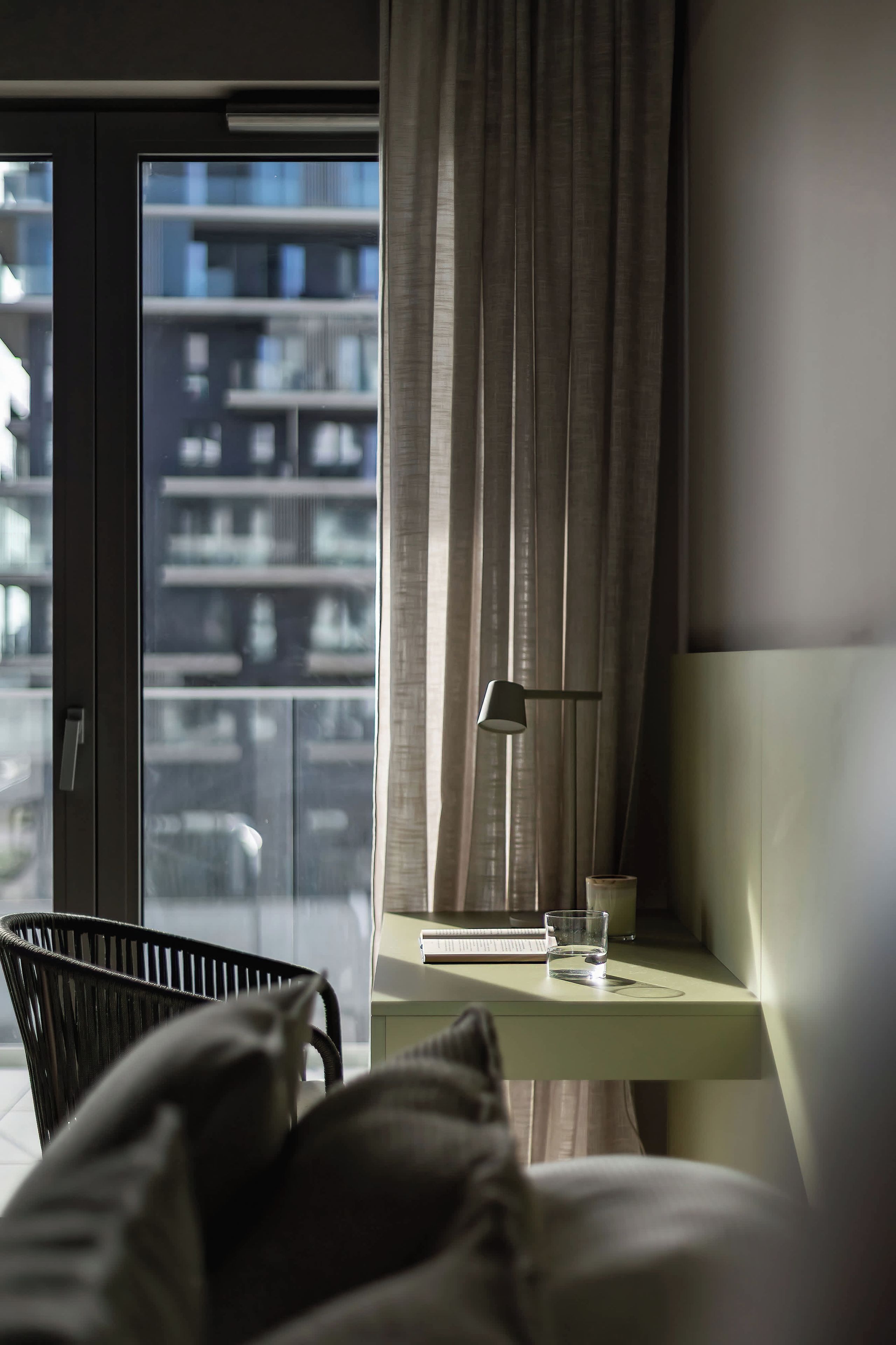 A hotel room with a bed, a desk with a lamp, a glass of water, and a view of a building through the window.