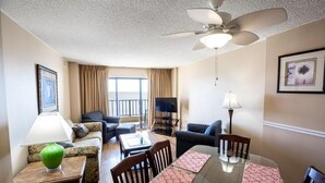 Quarto de hotel com mesa de jantar, cadeiras, sofá, televisão, ventoinha no teto e janela com cortinas.