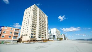 Un hotel frente a la playa con varios pisos, balcones y una playa de arena al fondo.