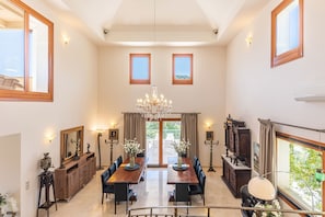 A spacious dining area with a chandelier, two windows, and a view of the outdoors. - Aeonia Grand Villa, with Pool & Walk to the Beach (Heraklion)