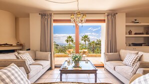 Un salon avec deux canapés, une table basse et un lustre. Une grande fenêtre offre une vue sur les palmiers, et il y a un balcon.