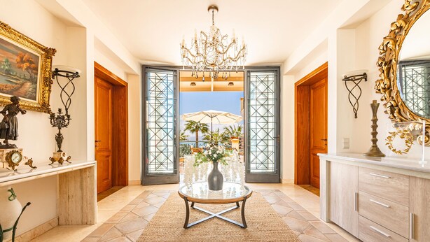 Un vaste hall d’entrée agrémenté d’un lustre, d’une table ronde surmontée d’un vase, et offrant une vue sur l’extérieur grâce à de grandes portes.