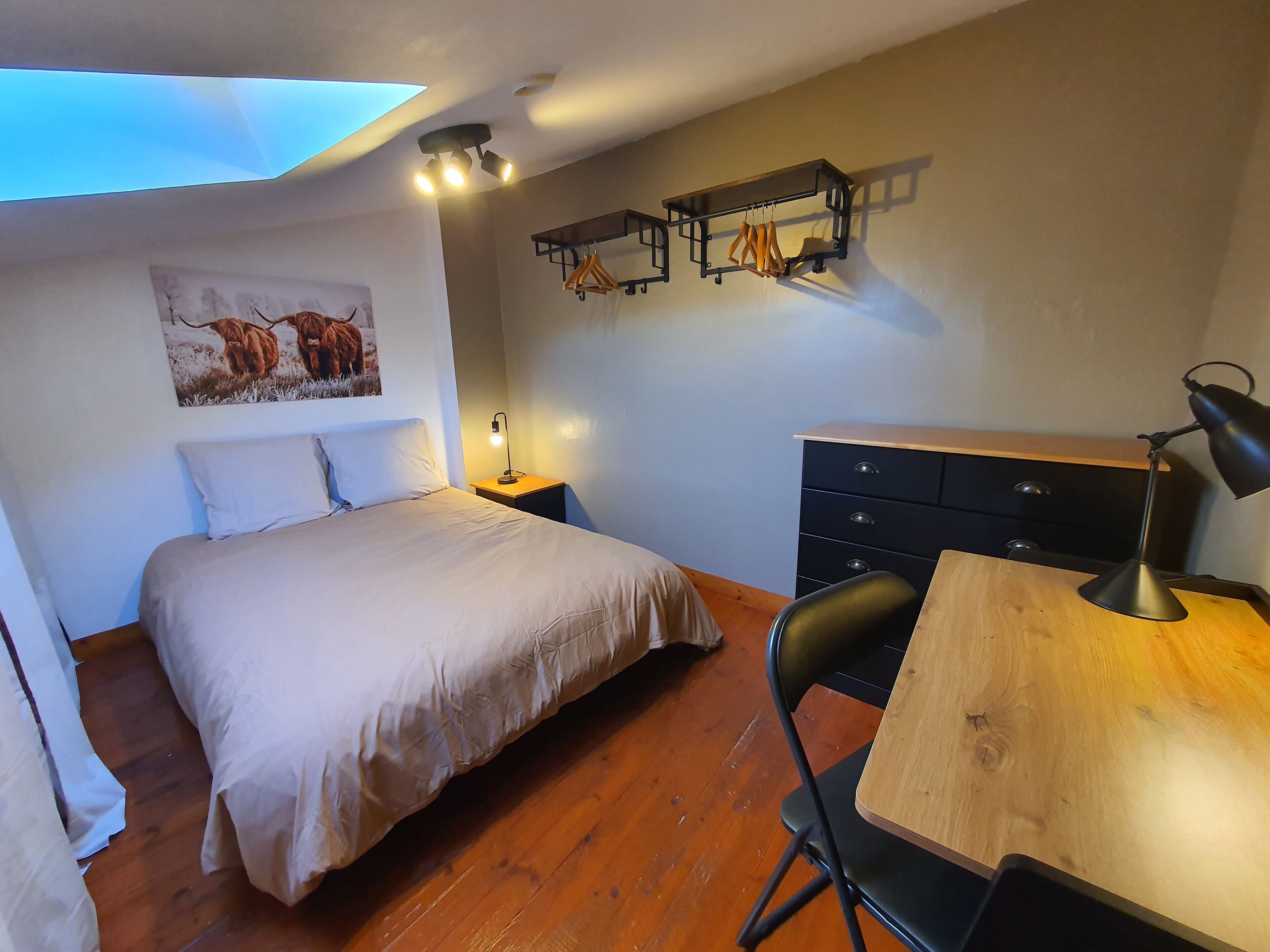 A bedroom with a bed, bedside table, desk, chair, and a wall-mounted shelf with clothes.