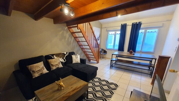 Ein Wohnzimmer mit einem schwarzen Ecksofa, einem Holztisch, einem gemusterten Teppich, einem Esstisch mit Bänken und einer Treppe.