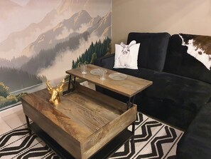 A living room with a wooden coffee table, a black sofa, and a mountain landscape mural on the wall. - Immersion at Le Trappeur Atypique Vendée-At the foot of the Cathedral-With parking (Luçon)