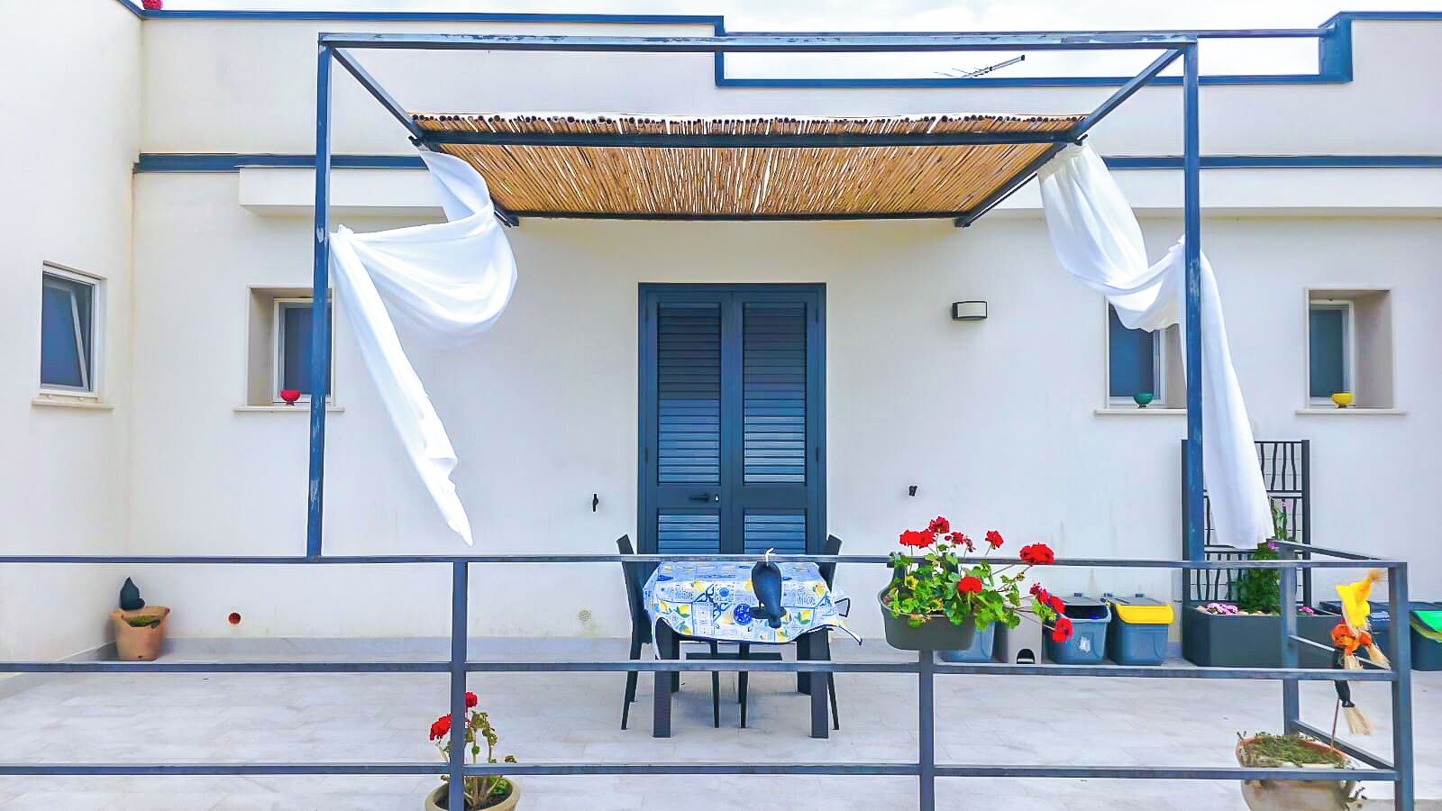 Un balcón con una mesa, sillas, plantas en macetas y una puerta azul.