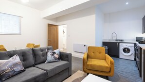 Een moderne woonkamer met een grijze bank, een gele fauteuil en een witte salontafel. In de achtergrond is een keuken met een wasmachine en een spoelbak zichtbaar.