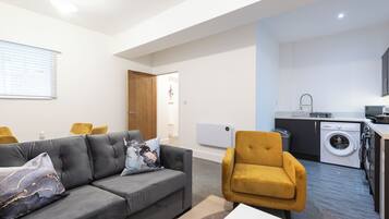 A modern living room with a grey sofa, a yellow armchair, and a white coffee table. A kitchen with a washing machine and a sink is visible in the background.