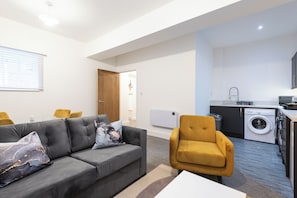 A modern living room with a grey sofa, a yellow armchair, and a white coffee table. A kitchen with a washing machine and a sink is visible in the background. - Old Dutch Embassy by Guestz (Hull)