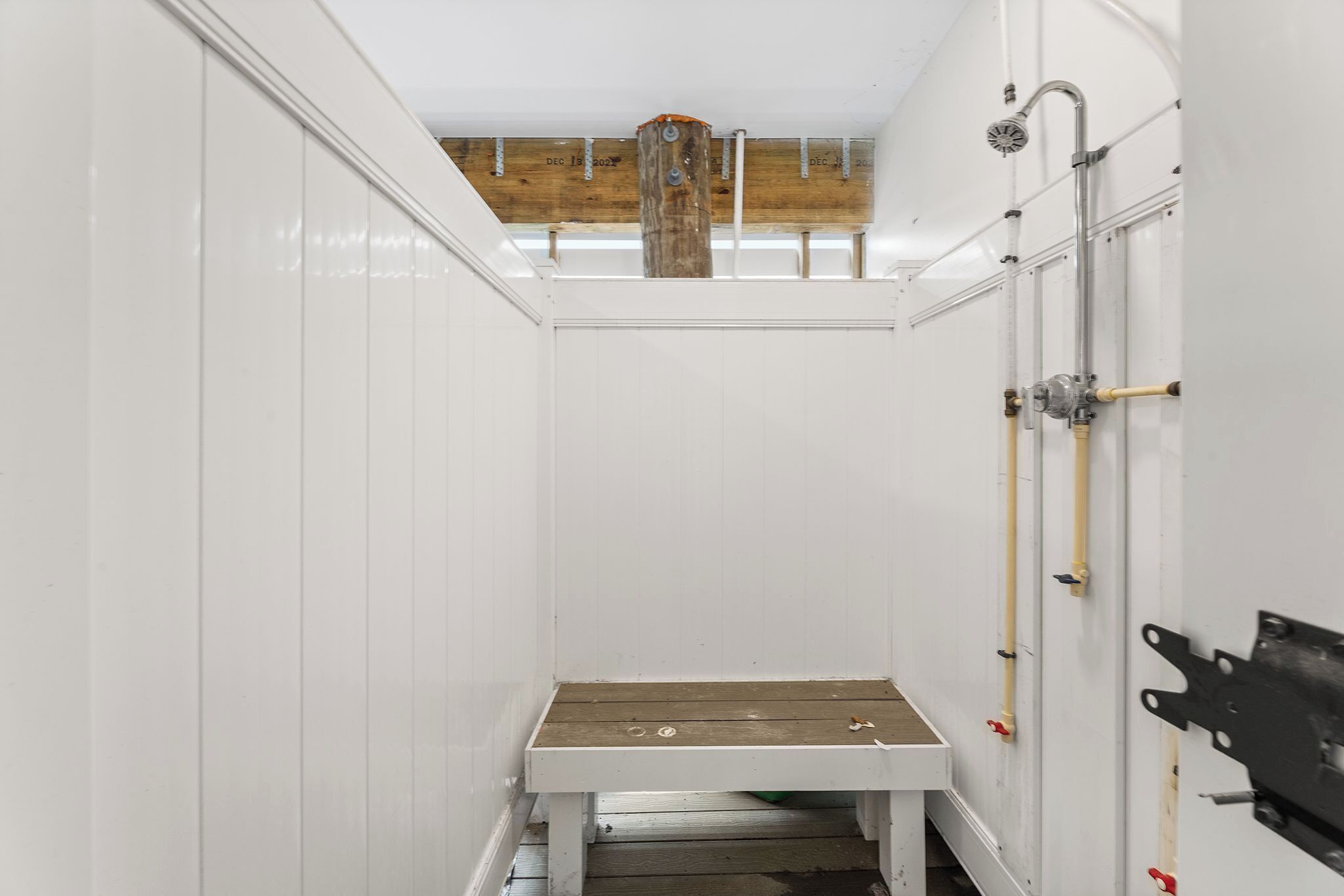 A narrow, white-walled room with a wooden bench, a visible water pipe, and a wooden beam ceiling.