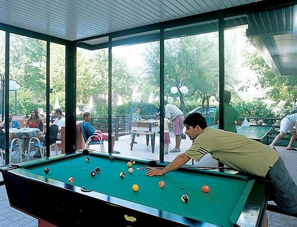 Une table de billard se trouve dans un espace extérieur abrité, d’où l’on peut admirer les personnes et la verdure environnante.