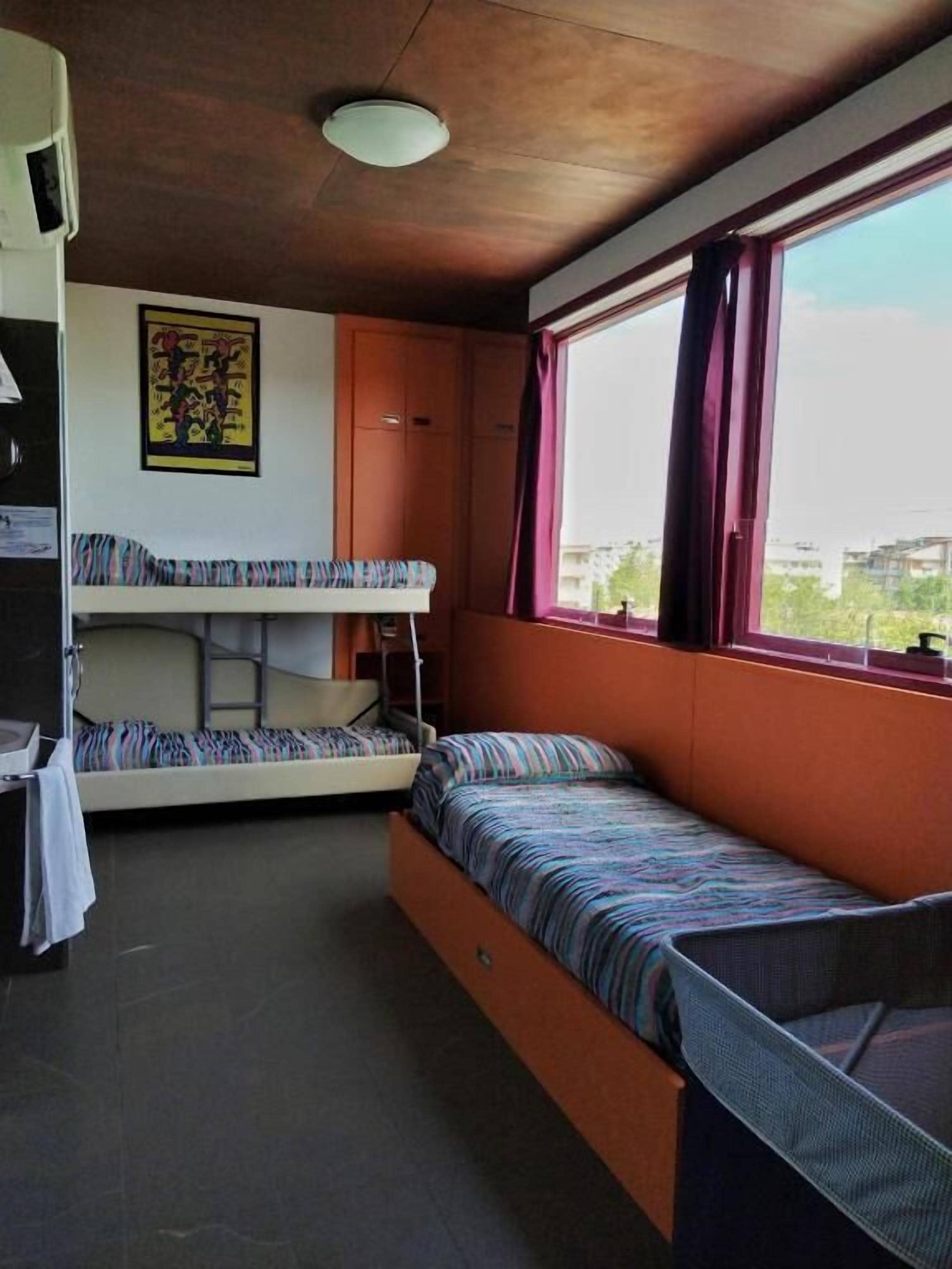 A dormitory room with bunk beds, a ceiling fan, a window with curtains, and a poster on the wall.