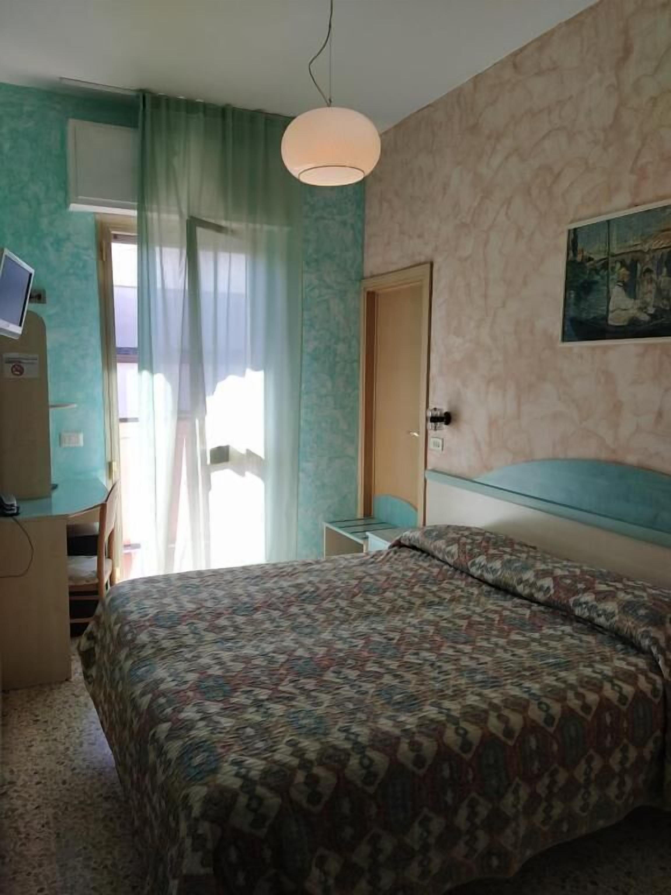 Un dormitorio con cama, escritorio con computadora, silla, puerta y ventana con cortinas.